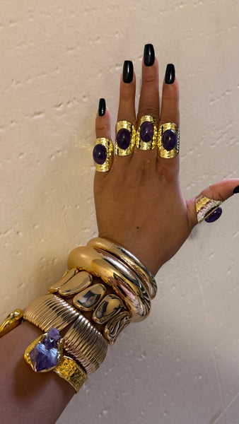 Hand wearing multiple gold rings with purple stones against a neutral background